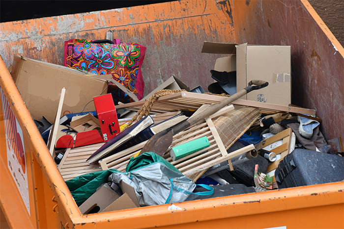 Ein orangefarbener Container gefüllt mit gemischtem Sperrmüll wie Holzlatten, Kartons, Haushaltsgegenständen und einer bunten Tasche, zur Entsorgung und zum Recycling vorbereitet.