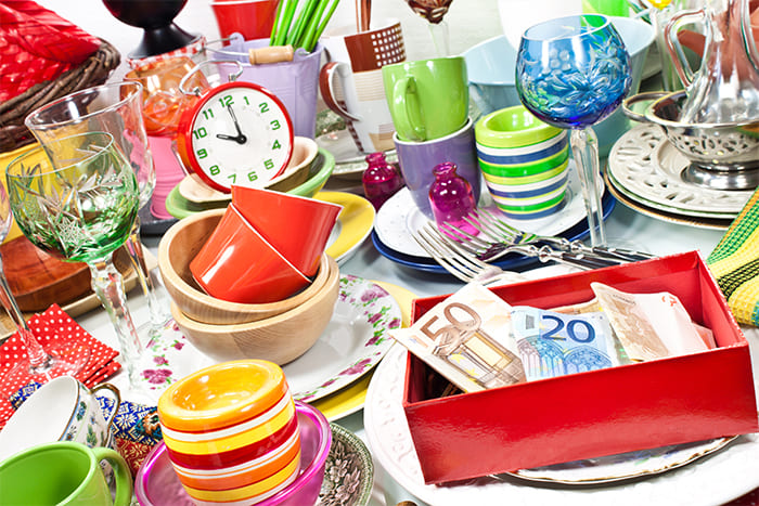 Verschiedene Gegenstände wie buntes Geschirr, Gläser, eine Uhr und eine rote Box mit Geldscheinen, die bei einer Nachlassauflösung gesichtet und sortiert werden.