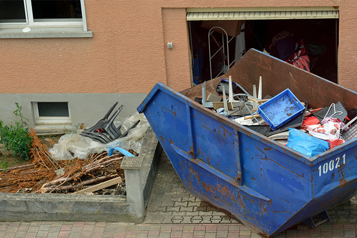 Blauer Abfallcontainer vor einer Garage, gefüllt mit verschiedenen Gegenständen und Baumaterialien, im Rahmen einer privaten Entrümpelung eines Hauses.