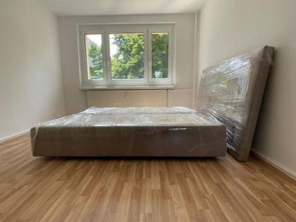 Ein Bettgestell und Kopfteil, sorgfältig mit transparenter Schutzfolie verpackt, in einem leeren Zimmer mit Laminatboden