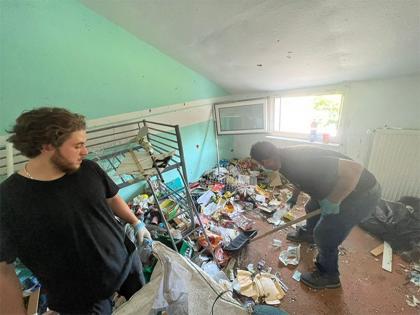 Zwei Personen, die in einer Messie-Wohnung Müll und Gegenstände entfernen, während der Raum für eine gründliche Entrümpelung vorbereitet wird