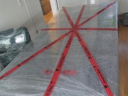 Ein großer Tisch, sicher mit Schutzfolie und rotem „Vorsicht Glas“-Klebeband für den Transport verpackt