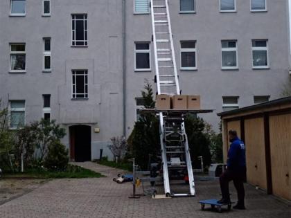 Ein Umzugsaufzug, der Umzugskartons aus einem oberen Stockwerk vor einem Mehrfamilienhaus transportiert, mit einem Mitarbeiter, der den Vorgang überwacht